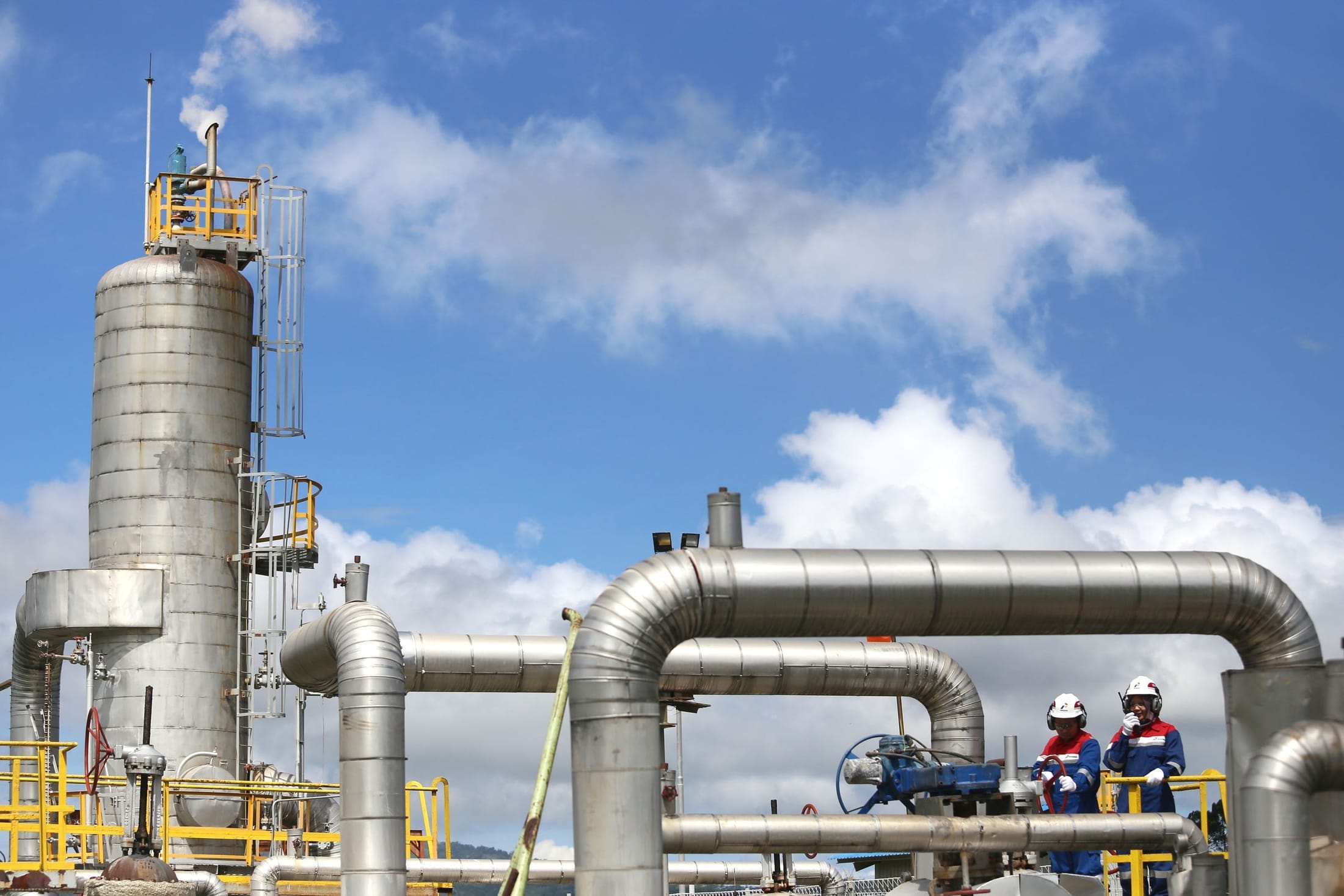 Two workers in protective equipment beside large insulated process piping